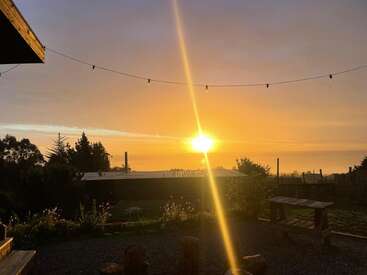 A warm sunset glows over a peaceful garden with string lights, a wooden bench, and lush greenery. Calm atmosphere, soft sky, and golden sunlight beams.