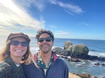 A smiling couple in sunglasses poses by the rocky ocean shore under a bright blue sky with scattered clouds. The sea looks calm and inviting.