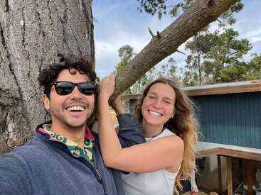 A smiling man wearing sunglasses and a woman with long hair stand beside a large tree outdoors. Both look happy, enjoying a sunny, nature-filled day together.