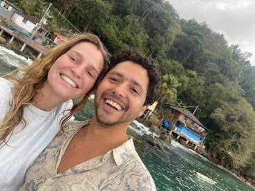 A smiling couple stands close together near a waterfront. Behind them are lush green trees, rustic houses on stilts, rocks, and gently flowing water. Vacation vibe.