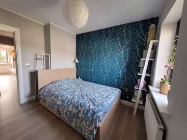 Esse quarto aconchegante tem uma cama de madeira com um edredom azul estampado, uma parede azul bem chamativa com galhos brancos, prateleiras, plantas e muita luz natural.