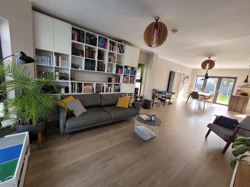 Uma sala de estar moderna com piso de madeira, sofá cinza, estantes de livros, plantas verdes e luz natural. Área de jantar e cozinha à vista, decoradas em estilo minimalista.