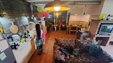 Quatro pessoas estão em uma cozinha aconchegante, decorada com balões coloridos. Elas preparam a comida, conversam e relaxam. O espaço tem um sofá estampado, decoração rústica e iluminação aconchegante.