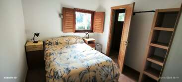 Um quarto aconchegante com uma cama de casal com roupa de cama floral, duas mesinhas de cabeceira de madeira, luminárias, uma janela com persianas, prateleiras abertas e uma porta de banheiro aberta.