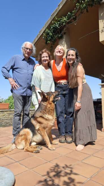 Four people and a German Shepherd pose outdoors on a sunny day. They stand on a terrace, smiling warmly, with a blue sky and house in the background.
