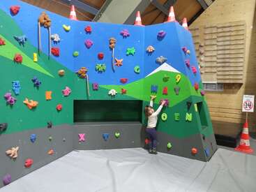 A young child climbs a colorful indoor bouldering wall decorated with animal shapes, letters, and numbers. Safety mats and traffic cones enhance the playful, safe environment.