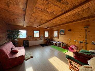 This cozy, sunlit room features wooden walls and ceiling, colorful couches, a green yoga mat, playful decor, indoor plants, and three bright windows with mountain views.