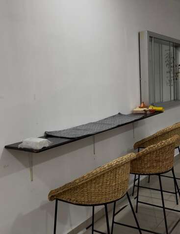 This image shows a minimalist setup with a long black wall-mounted shelf, two woven chairs, some items on the shelf, and a window with glass panes.