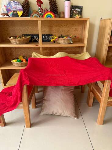 Two wooden chairs are draped with red and yellow cloths, forming a play fort. Behind, shelves hold baskets, books, pinecones, and colorful wooden toys. Cozy, inviting scene.
