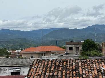 Red-tiled roofs in the foreground, modern and rustic buildings, with lush green mountains and cloudy sky backdrop, create a tranquil and picturesque town scene.