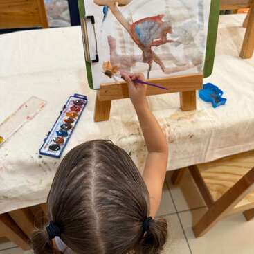 A young child with pigtails is painting abstract art using watercolors at a small easel. Brushes, a color palette, and art supplies are nearby.