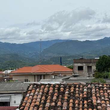 Red-tiled roofs in the foreground, modern and rustic buildings, with lush green mountains and cloudy sky backdrop, create a tranquil and picturesque town scene.