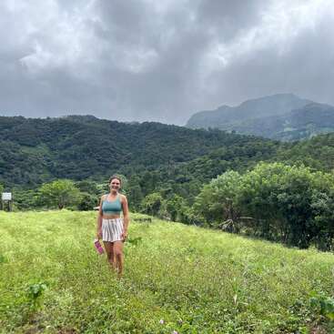 The image depicts a woman standing in a lush grassy field, set against a backdrop of trees and mountains under an overcast sky.