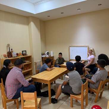 A small classroom with children and a teacher seated around wooden tables. The teacher is guiding a lesson using a whiteboard, fostering interactive learning.