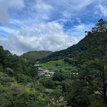 The image depicts a serene mountainous landscape with lush greenery, featuring a small village nestled among trees and hills, set against a blue sky with white clouds.