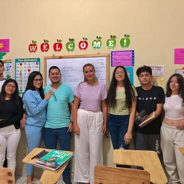 The image shows a group of students and a teacher standing in front of a whiteboard and colorful wall decorations in a classroom setting.