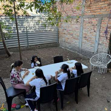 Five girls sit around a white table outdoors, eating sandwiches. Trees, a brick wall, and hanging chair create a shaded, relaxed backyard atmosphere. Toys lie nearby.