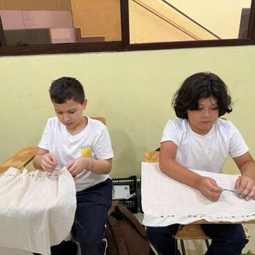 Duas crianças, sentadas em carteiras, estão concentradas costurando um tecido branco. Ambas vestem camisetas brancas e estão sentadas em uma sala de aula, com uma janela atrás delas.
