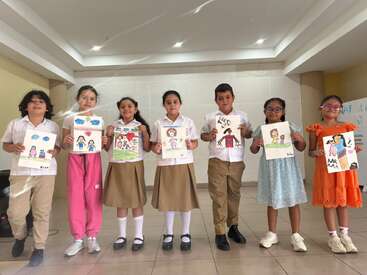 Seven children stand in a row indoors, proudly holding colorful drawings. They wear school uniforms or casual clothes and smile, presenting their artwork to the camera.