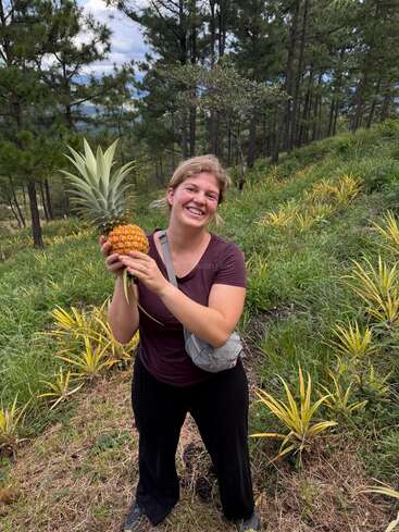 A smiling woman stands outdoors on a hillside farm, holding a freshly harvested pineapple, surrounded by green grass, young pineapple plants, and tall pine trees.