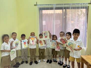 A group of young children, dressed in school uniforms, proudly display colorful drawings they created, standing inside a brightly lit classroom with a large window.