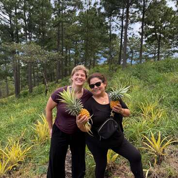 Duas mulheres sorriem alegremente, segurando abacaxis frescos em um campo verdejante. Cercadas por pinheiros altos e pés de abacaxi, elas curtem a natureza juntas.