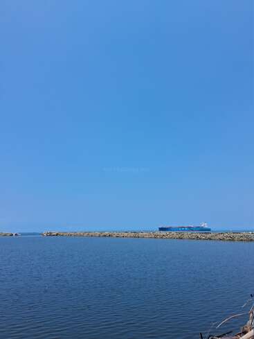 Un grand cargo bleu vogue à l'horizon, avec au premier plan une mer calme et un brise-lames en pierre, sous un ciel bleu clair et lumineux. Une scène paisible.