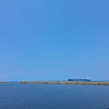 A large blue cargo ship sails on the distant horizon, calm water in the foreground, stone breakwater, under a clear, bright blue sky. Peaceful scene.