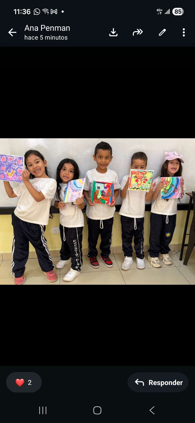 Cinco niños felices están en fila, sosteniendo sus coloridos dibujos. Visten uniformes a juego y sonríen orgullosos en un aula con una pizarra blanca detrás de ellos.