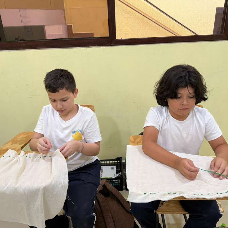 Two children, sitting at desks, are focused on sewing white fabric. Both wear white t-shirts and sit in a classroom environment with a window behind them.