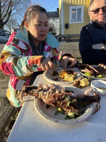 A woman in a colorful jacket enjoys a meal outdoors with another person. Plates hold grilled meat, vegetables, and corn, set on a white table in sunlight.