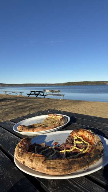 Dois pratos de pizza meio comidos estão sobre uma mesa de piquenique de madeira à beira de um lago tranquilo, sob um céu azul claro, com bancos ao fundo.