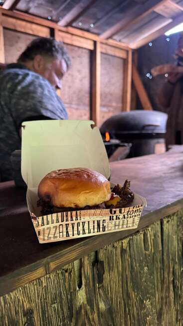 Um sanduíche de carne desfiada recém-preparado está dentro de uma caixa sobre um balcão rústico de madeira. Ao fundo, um homem trabalha perto de um forno a lenha.