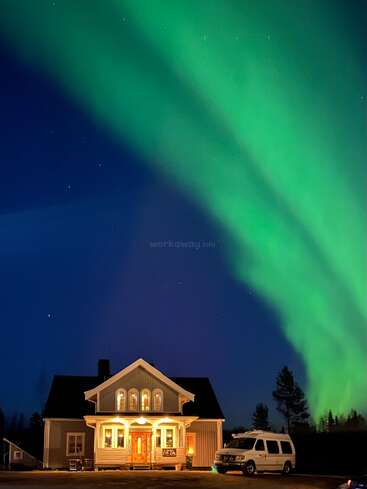 Uma casa aconchegante e bem iluminada ergue-se sob um céu noturno escuro, iluminado pela aurora boreal de um verde vibrante. Uma van branca está estacionada ao lado da casa, com as árvores em silhueta.