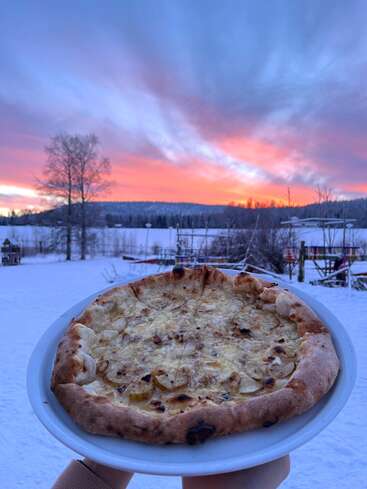 Uma pizza recém-assada em um prato branco é retratada ao ar livre, tendo como pano de fundo uma paisagem nevada, com um pôr do sol colorido deslumbrante e nuvens em tons pastéis iluminando o céu.