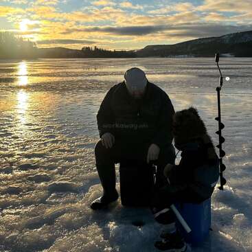 Um homem e uma criança estão sentados num lago congelado ao pôr do sol, pescando no gelo juntos. Uma broca está ali perto, rodeada por neve cintilante e uma paisagem serena.