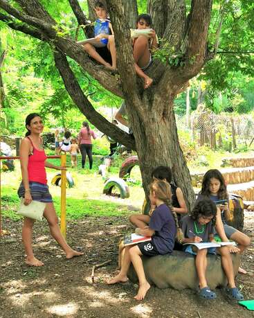 As crianças estão lendo e escrevendo ao ar livre, algumas sentadas em uma árvore, outras em um pneu, enquanto um adulto sorridente está por perto. O ambiente é exuberante e verde.