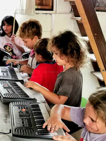 Quatro crianças tocam com entusiasmo em pequenos teclados, enquanto outra criança toca bateria ao fundo, todas imersas em fazer música juntas em um ambiente interno aconchegante.