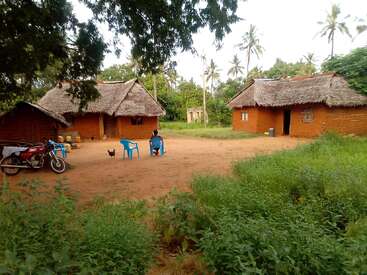 The image depicts a serene rural scene with two thatched-roof houses, a dirt courtyard, and a motorcycle, surrounded by lush greenery and palm trees.
