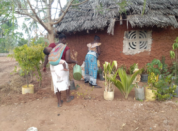 The image depicts two women and a baby in front of a hut, with one woman carrying the baby and the other watering plants.