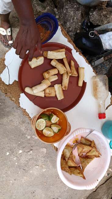 This image shows a hand reaching for cassava pieces on a red tray, with samosas, lime slices, and some condiments arranged on a rough outdoor surface.