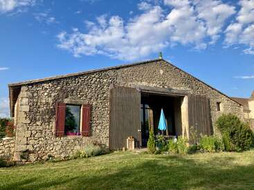 This image shows a rustic stone barn with wooden doors, a lush green yard, scattered plants, a blue umbrella, and a bright sky with scattered clouds.