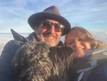 A couple smiles for a selfie with their fluffy black dog, outdoors on a sunny day. They look happy, surrounded by sky and clouds behind them.