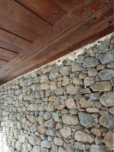 Das Bild zeigt eine Nahaufnahme einer rustikalen Steinmauer, über der sich eine Holzdecke mit sichtbarer Maserung und verwitterter Oberfläche befindet. Natürliche, erdige Ästhetik.
