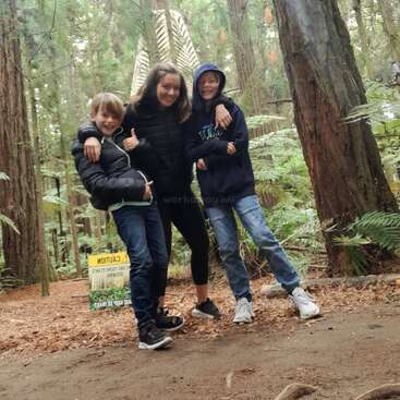 La imagen muestra a tres niños posando en una zona boscosa, dos de ellos de pie delante de una mujer y otro al fondo, todos con atuendo informal.
