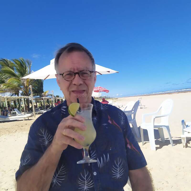 The image depicts a man standing on a sandy beach, holding a cocktail with a lime wedge, wearing a dark blue Hawaiian shirt with white palm fronds, set against a bright blue sky.