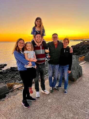 Una feliz familia de seis miembros sonríe alegremente cerca de una costa rocosa al atardecer, disfrutando de la compañía mutua con el cielo dorado y el mar en calma a sus espaldas.