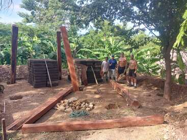 Quatre hommes se tiennent sur un chantier de construction avec de grandes poutres en bois, des briques empilées et quelques outils, entourés de verdure et d'arbres, travaillant à l'ombre.