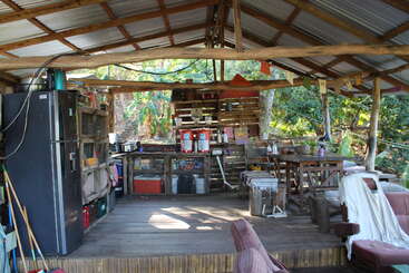 Cette cuisine rustique en plein air comprend une terrasse en bois, des étagères ouvertes, un réfrigérateur, des tables avec des chaises et divers ustensiles. Entourée d'une végétation luxuriante, sous un toit en tôle.