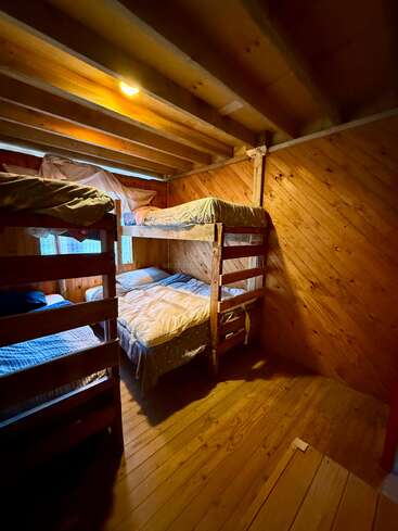 Esse quarto aconchegante de madeira apresenta dois conjuntos de beliches com roupas de cama simples, iluminação suave de uma única lâmpada e paredes de madeira quente, criando uma atmosfera rústica.